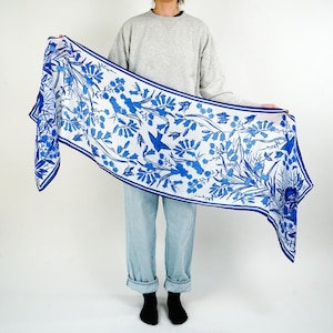 May include: A long, rectangular scarf with a white background and a blue floral and bird pattern. The scarf is held up, displaying the intricate design. The edges are trimmed with a solid blue border. The scarf appears to be made of a lightweight fabric.