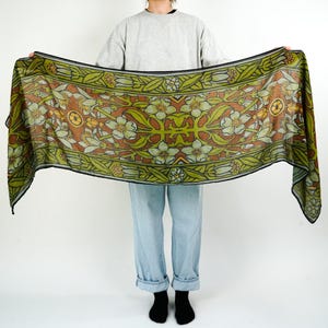 May include: A rectangular scarf with a floral pattern in shades of green, brown, and white. The scarf is held up, displaying its full length and design. The edges are outlined in black.