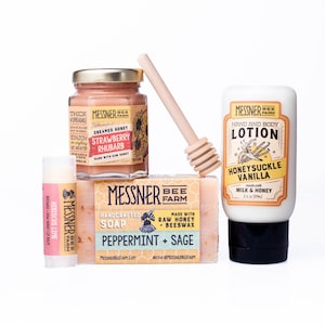 May include: A collection of Messner Bee Farm products, including a jar of strawberry rhubarb creamed honey, a bar of peppermint and sage soap, a honeysuckle vanilla lotion, a hibiscus lime lip balm, and a wooden honey dipper. The products are arranged on a white surface.
