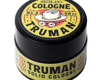 May include: A small black circular container of Truman solid cologne. The lid features a yellow label with a graphic of a white fedora hat and round glasses, with the text SOLID COLOGNE, No. 2, .50 oz, and TRUMAN printed in black.