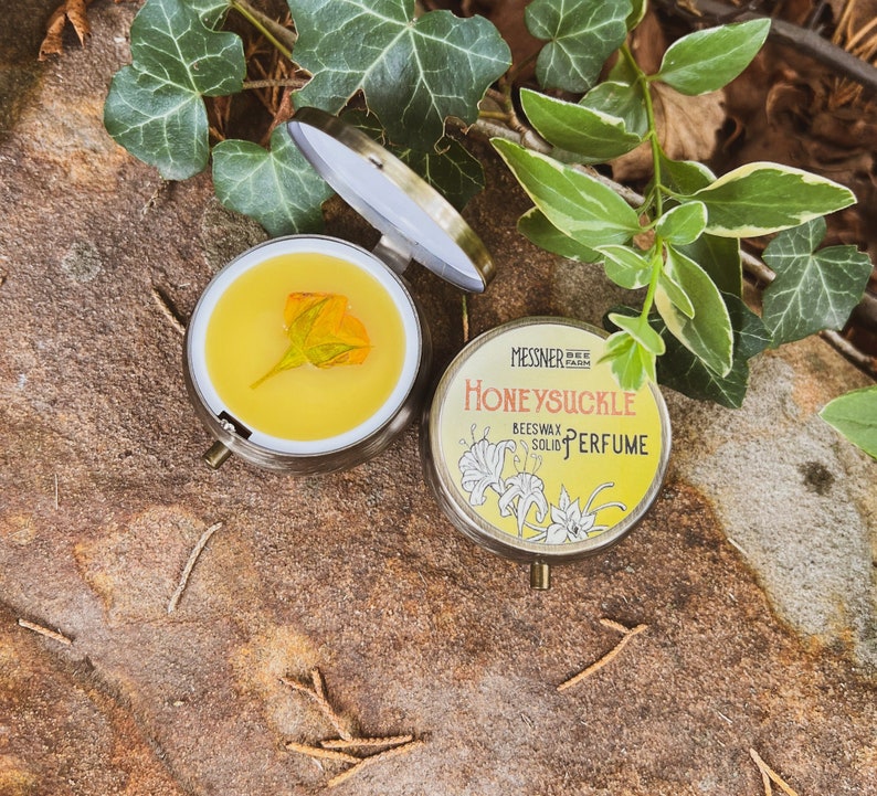 May include: A round tin container with a gold lid and a floral design. The label reads "MESSNER FARM BEESWAX SOLID PERFUME HONEYSUCKLE". The tin is open and a small amount of yellow beeswax is visible inside.