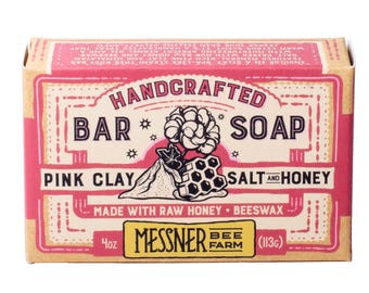 May include: A rectangular bar of Messner Bee Farm handcrafted soap in a tan and pink cardboard box. The label features illustrations of a flower, a pile of salt, and a honeycomb, with text reading Pink Clay, Salt and Honey, 4oz, and 113g.