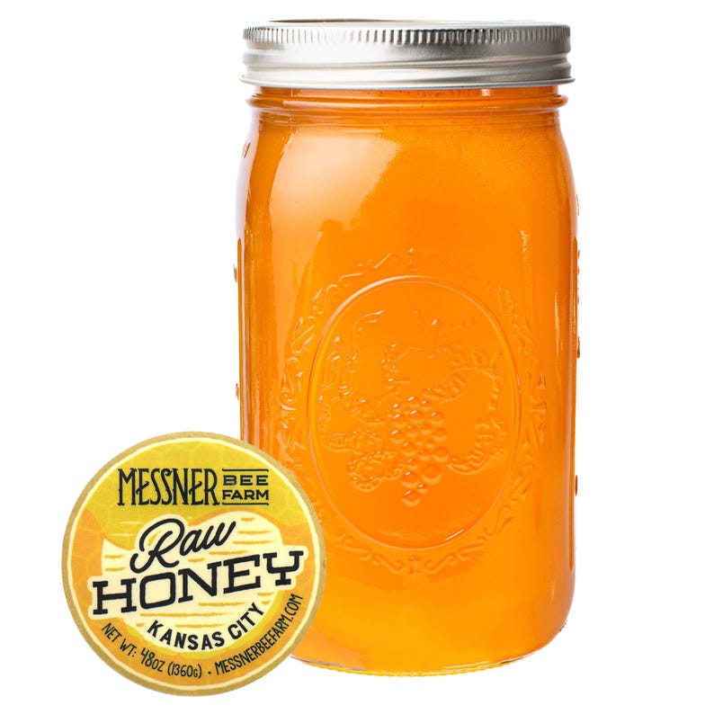 May include: A clear glass jar filled with golden-colored raw honey. The jar has a silver lid and embossed detailing. A round label with the text "Messner Bee Farm Raw Honey Kansas City" is next to the jar. The net weight is 48oz.