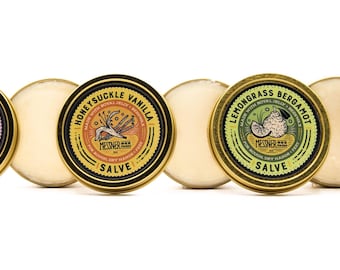 Op de afbeelding: Vier ronde blikken met zalf met gouden deksels en kleurrijke etiketten. De etiketten bevatten illustraties van bloemen, kruiden en tekst die de geur van elke zalf beschrijft: Lavendel Havermout, Kamperfoelie Vanille, Citroengras Bergamot en Puur en Simpel.