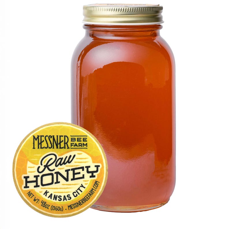 May include: A clear glass jar filled with raw honey, with a gold-colored lid. A round yellow label with the text "Messner Bee Farm Raw Honey Kansas City" is on the left. The jar contains 48 oz of honey.