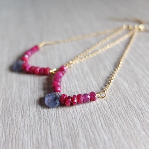 May include: Delicate gold chain earrings. Each earring features a curved line of small, bright pink gemstones, with a single, larger, blue-grey stone. The design is elegant and adds a touch of colour.