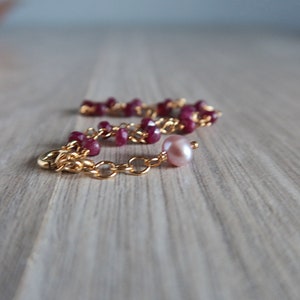Op de afbeelding: Een gouden kettingarmband met rode edelsteen kralen en een enkele roze parel bedel.