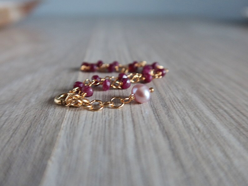 Op de afbeelding: Een gouden kettingarmband met rode edelsteen kralen en een enkele roze parel bedel.