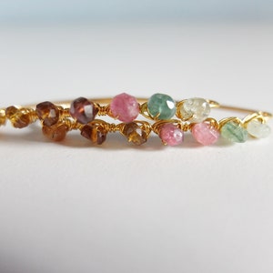 May include: A gold wire bracelet with a row of colorful gemstones. The bracelet features pink, green, and brown gemstones.