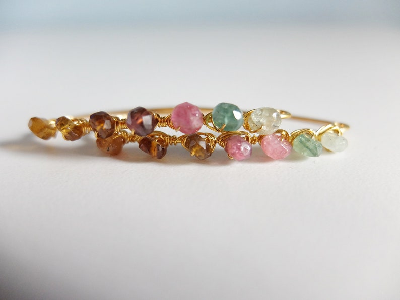 May include: A gold wire bracelet with a row of colorful gemstones. The bracelet features pink, green, and brown gemstones.