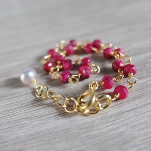 Op de afbeelding: Een gouden kettingarmband met een enkele witte parelbedel en meerdere kleine, gefacetteerde, robijnrode kralen.