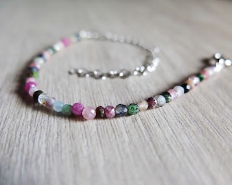 May include: A delicate bracelet featuring a strand of colorful, faceted gemstones in shades of pink, green, blue, and clear. The beads are strung on a silver chain, with a clasp closure. The bracelet is displayed on a light wood surface.