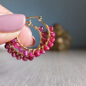 Op de afbeelding: Gouden oorringen versierd met levendige, dieproze robijnkralen. De kralen zijn langs de omtrek van de oorring geregen, waardoor een textuurrijk, opvallend ontwerp ontstaat. De achtergrond is zacht en wazig.