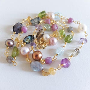 Puede incluir: Un collar de piedras preciosas multicolores con una variedad de cuentas moradas, verdes, amarillas y perlas. Las cuentas están ensartadas en una delicada cadena dorada. Una hermosa joya.