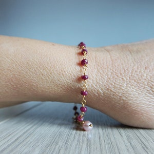 Op de afbeelding: Een gouden kettingarmband met kleine, donkerrode kralen en een enkele, grotere roze kraal aan het einde.