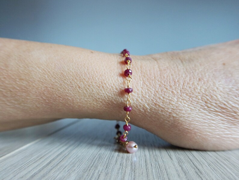 Op de afbeelding: Een gouden kettingarmband met kleine, donkerrode kralen en een enkele, grotere roze kraal aan het einde.