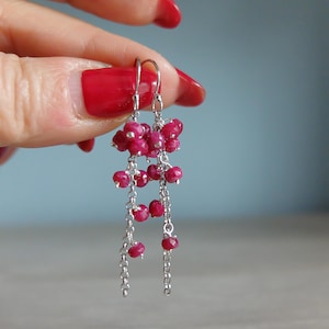 May include: Dangling earrings with clusters of small, round, deep red beads suspended from delicate silver chains. The earrings have a cascading design, with multiple strands of beads and chains. The background is a soft blue.