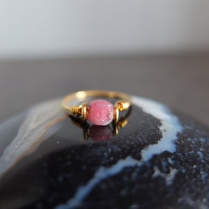 K&ouml;nnte beinhalten: Ein zarter Goldring mit einem rosafarbenen Edelstein. Der Ring liegt auf einem schwarzen und wei&szlig;en Achat-Stein.