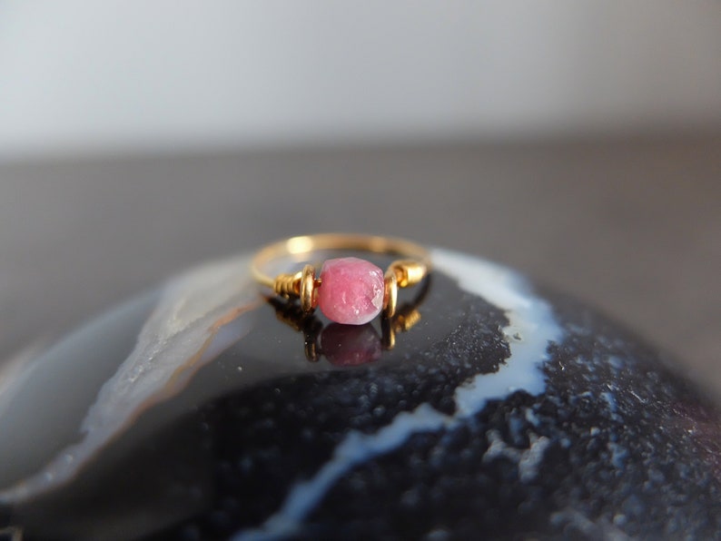 K&ouml;nnte beinhalten: Ein zarter Goldring mit einem rosafarbenen Edelstein. Der Ring liegt auf einem schwarzen und wei&szlig;en Achat-Stein.