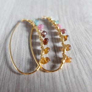 May include: A pair of gold hoop earrings with a row of colorful gemstones hanging from each hoop. The gemstones are in shades of pink, green, and brown.