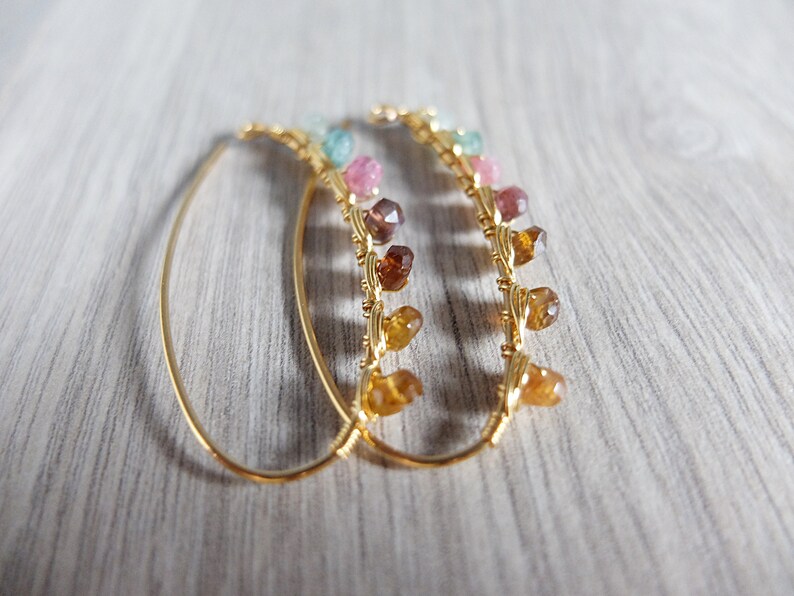 May include: A pair of gold hoop earrings with a row of colorful gemstones hanging from each hoop. The gemstones are in shades of pink, green, and brown.