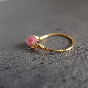 May include: A delicate gold ring with a pink gemstone set in the band. The ring is simple and elegant, perfect for everyday wear.