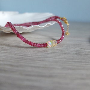 May include: A delicate pink gemstone necklace with three iridescent opal beads. The necklace is on a white seashell and a wooden surface.