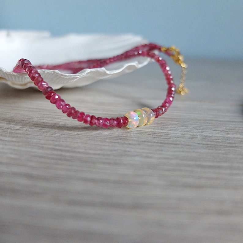 May include: A delicate pink gemstone necklace with three iridescent opal beads. The necklace is on a white seashell and a wooden surface.