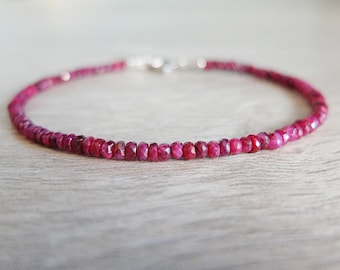 May include: A delicate bracelet composed of small, faceted, deep pinkish-red gemstone beads arranged in a single strand. The bracelet features a metallic clasp mechanism at the back, connecting the circular string of vibrant, polished stone beads.