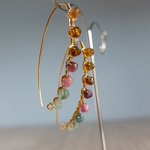 May include: Gold hoop earrings with a cascading row of colorful gemstones. The gemstones are in shades of pink, green, and brown.