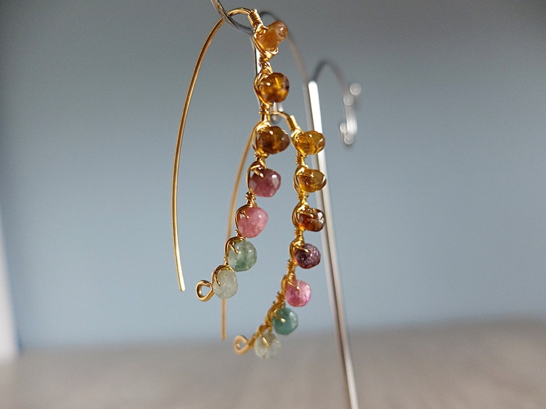 May include: Gold hoop earrings with a cascading row of colorful gemstones. The gemstones are in shades of pink, green, and brown.
