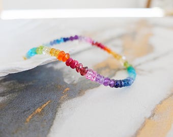 Op de afbeelding: Een delicate armband bestaande uit kleine, gefacetteerde, veelkleurige edelsteenkralen gerangschikt in een levendig regenboogverloop. De kralen gaan over in tinten rood, oranje, geel, groen, blauw en paars, wat een continu spectrum vormt.