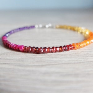 May include: A multi-colored gemstone bracelet with purple, pink, red, orange, and yellow beads. The bracelet is made with small, faceted beads and has a silver clasp.