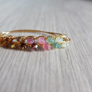 May include: A gold wire wrapped bracelet with a rainbow of gemstones. The bracelet features brown, pink, and green stones.