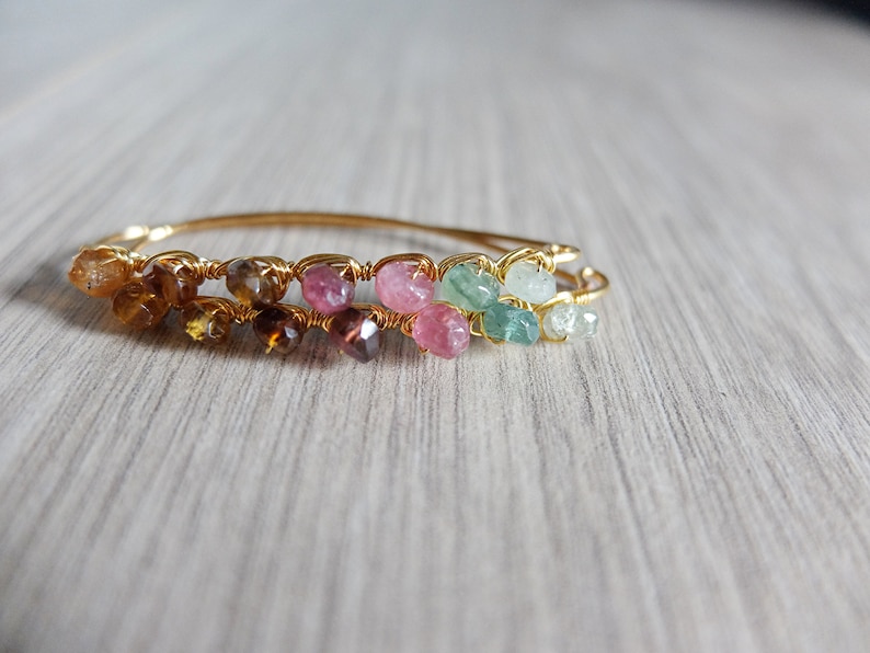May include: A gold wire wrapped bracelet with a rainbow of gemstones. The bracelet features brown, pink, and green stones.