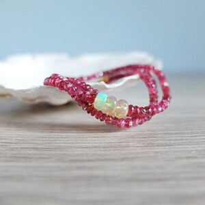 May include: A delicate pink gemstone necklace with three iridescent opal beads. The necklace is displayed on a white seashell.