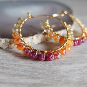 May include: Gold hoop earrings with a gradient of gemstones. The earrings feature small, faceted stones in shades of yellow, orange, and red.