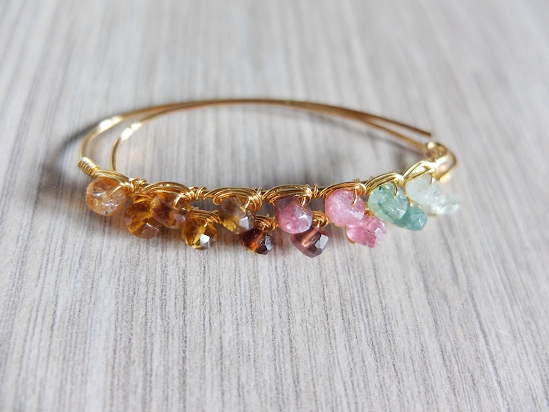 May include: A gold wire wrapped bracelet with a rainbow of gemstones. The bracelet features a variety of colors including brown, orange, pink, green, and blue.