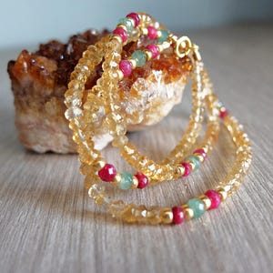 Puede incluir: Un brazalete de varias hebras con cuentas de citrino dorado, piedras preciosas rojas y verdes y cuentas de metal dorado. El brazalete se exhibe sobre una piedra rugosa de color marrón anaranjado.
