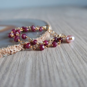 Op de afbeelding: Een delicate gouden kettingarmband met kleine rode edelsteen kralen en een enkele roze parel bedel.