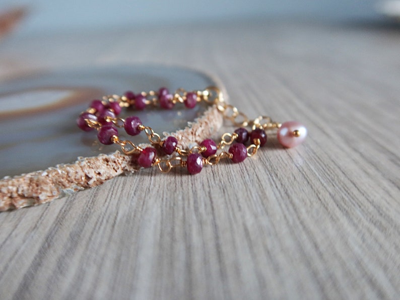 Op de afbeelding: Een delicate gouden kettingarmband met kleine rode edelsteen kralen en een enkele roze parel bedel.