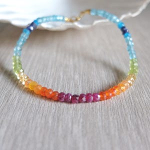 May include: A beaded bracelet featuring a rainbow of colors, including red, orange, yellow, green, blue, and purple. The bracelet is made of small, faceted beads strung together with a gold-colored clasp. The bracelet is displayed on a light gray surface.