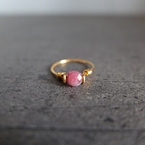 K&ouml;nnte beinhalten: Ein zarter Goldring mit einem einzigen rosafarbenen Edelstein. Der Ring ist aus Draht gefertigt, der um den Stein gewickelt ist.