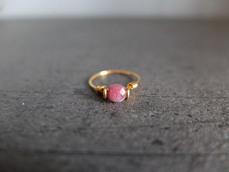 K&ouml;nnte beinhalten: Ein zarter Goldring mit einem einzigen rosafarbenen Edelstein. Der Ring ist aus Draht gefertigt, der um den Stein gewickelt ist.