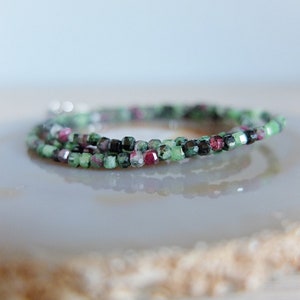 Op de afbeelding: Een delicate armband gemaakt met kleine, gefacetteerde kralen in tinten groen, roze en zwart. De kralen zijn geregen op een zilveren ketting.