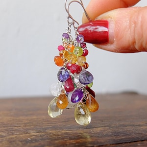 Op de afbeelding: Hangende oorbellen met een cluster van kleurrijke edelstenen. De oorbellen hebben verschillende vormen en kleuren, waaronder rood, oranje, geel, paars en groen. De oorbellen worden door een hand vastgehouden.