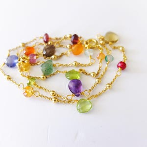May include: A multi-strand necklace with colorful gemstones and gold-colored chain. The gemstones are various shapes and colors, including purple, green, orange, and blue. The necklace is displayed on a white surface.