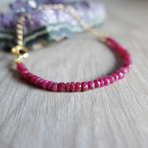 May include: A necklace with faceted, deep pink ruby beads on a delicate gold chain, secured with a gold clasp. The background includes a blurred purple crystal and a light wooden surface.