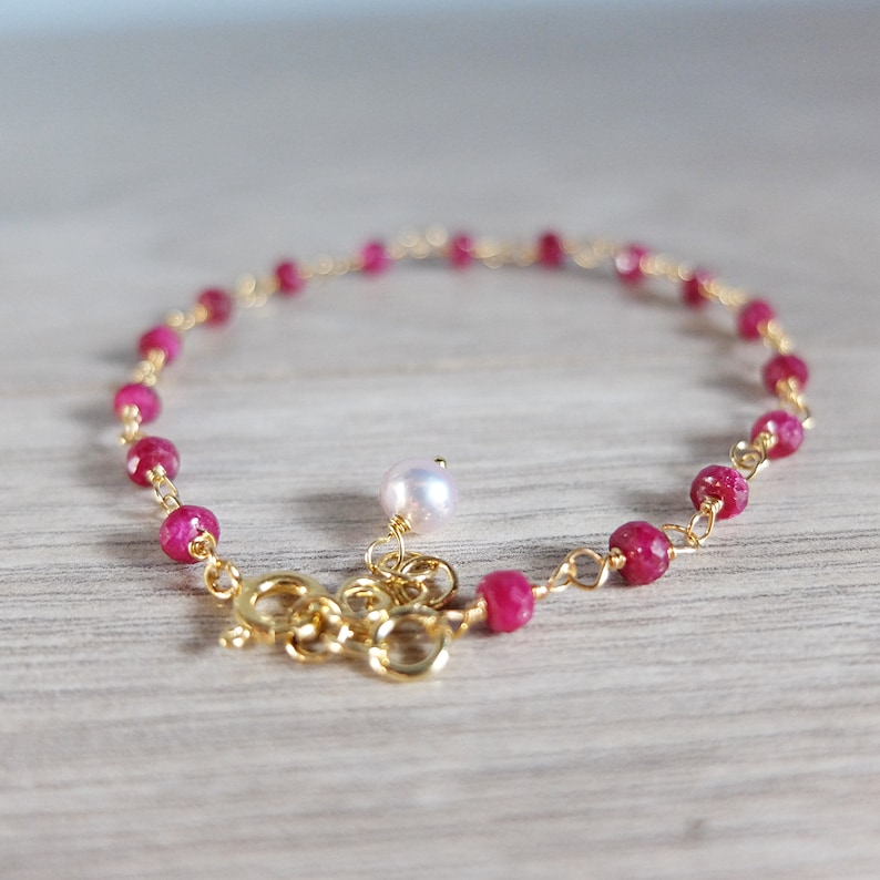 Op de afbeelding: Een delicate gouden kettingarmband met kleine rode edelsteen kralen en een enkele witte parel bedel.