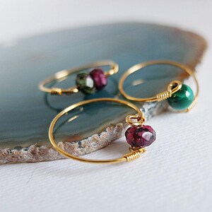 May include: Three delicate gold rings, each featuring a different gemstone. One ring has a green and burgundy stone, another has a green stone, and the third has a burgundy stone. The rings are displayed on a blue and brown surface.